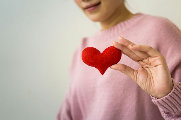 Content unrecognizable female wearing pink sweater standing on white background with small handmade heart in hand in light room inside